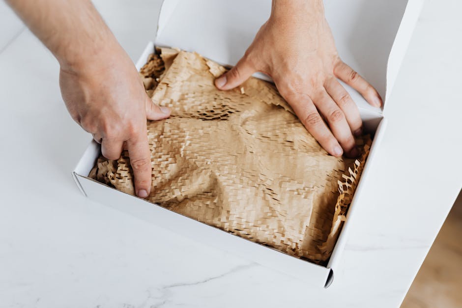 flexible packaging vs rigid packaging - From above crop adult male packing ordered product or gift in white cardboard box with craft paper for safety postal delivery at white marble table