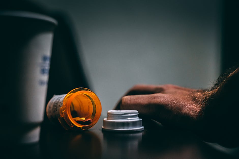 pharmaceutical packaging requirements - Moodily lit scene of an open prescription bottle and hand on a table, depicting healthcare themes.